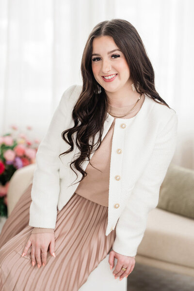 Karli Dawson, second photographer and content creator at Kari Dawson + Co, seated on a neutral-toned couch in a soft, feminine setting, wearing a cream jacket and blush dress