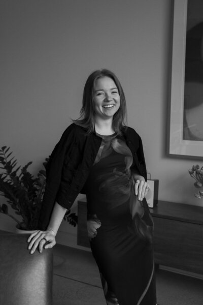 Documentary wedding photographer, Eryndae, smiling at camera with one hand on hip and one hand leaning on a leather chair