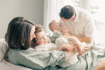 Mother and father sitting and tickling their two young children during an in-home lifestyle session in Kansas City, captured in soft natural light.
