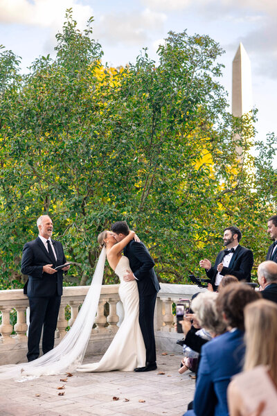 A couple shares a kiss during an outdoor wedding ceremony. They stand on a terrace surrounded by greenery, with guests seated nearby. The bride wears a white gown and veil, while the groom is in a dark suit. A tall monument is visible in the background.