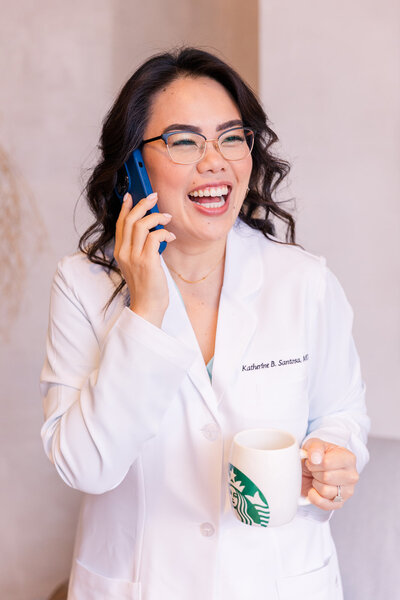 Woman doctor in white coat standing and laughing on the phone while holding a Starbucks coffee mug Laure Photography brand session