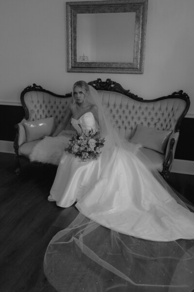 A black and white photograph of a bride sitting on an ornate, tufted vintage sofa. She is wearing a strapless white wedding gown with a full skirt and a long train that pools on the floor, while holding a bridal bouquet in her lap.