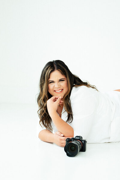 Adriana Montoya Photo lying on the ground, holding her camera close to her face. She is looking at the camera (not the one she is holding) and smiling.