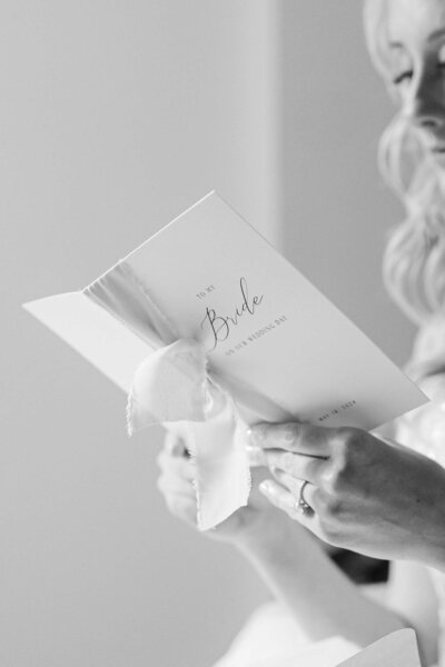 Black and white close-up of a bride reading a heartfelt letter on her wedding day. The card, tied with a soft fabric ribbon, reads “To my Bride on our Wedding Day” with the date elegantly printed below. The bride’s engagement ring is visible as she gently holds the note, capturing an emotional and intimate pre-ceremony moment.