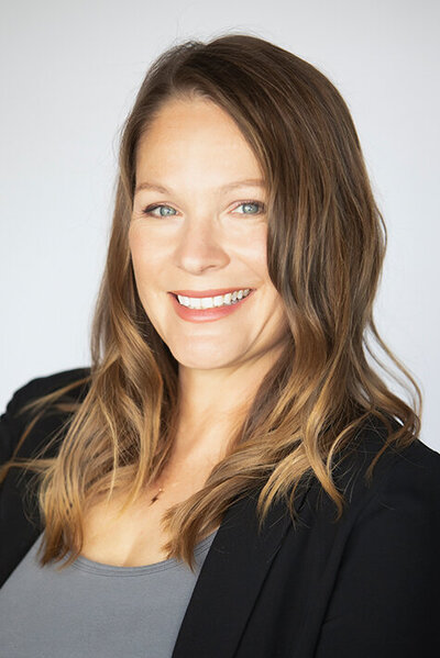 Headshot of a woman with light brown hair, light eyes, a black jacket and grey shirt.
