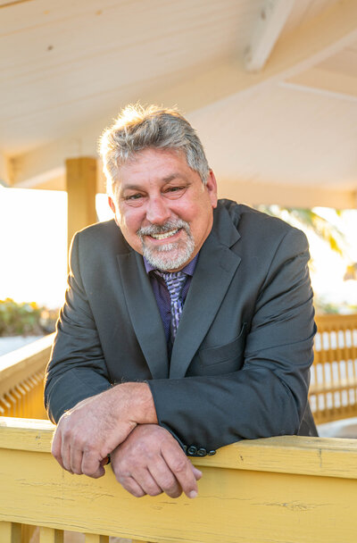 Photo of John Corbenllini smiling in a grey suit leaning against a wooden railing in an outdoor gazebo on the beach