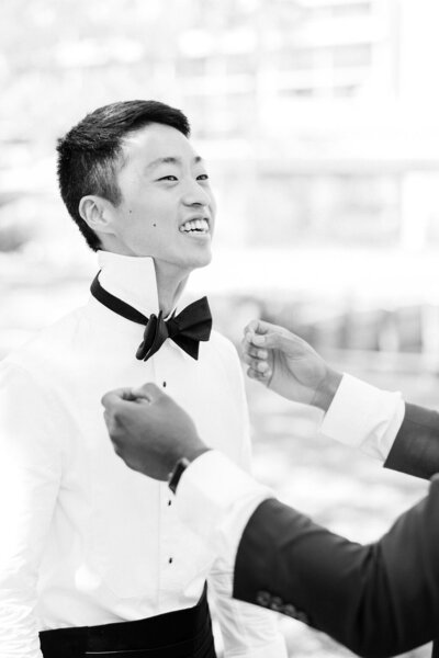 Groom beams while a friend straightens his black bow tie during the getting-ready moments of a Hilton Head Island wedding—captured by luxury wedding photographer Amia Marcell.