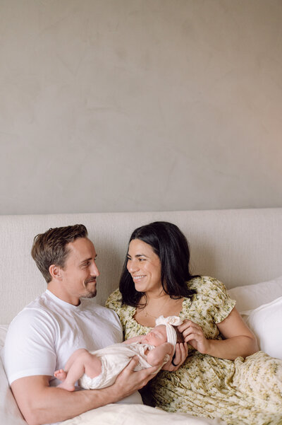 A couple sits together on their bed smiling at each other while holding their newborn baby, captured by a Northern Virginia family photographer during an in-home session.