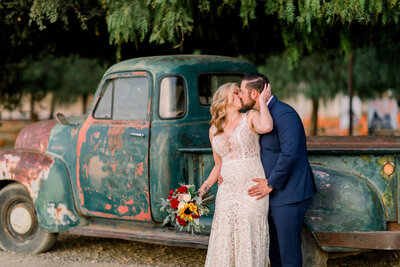 Bride turns back and shares a kiss with her new husband as he holds her from behind while standing next to an antique green truck