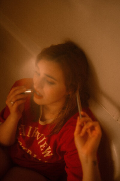 A woman in a red Fairview tee-shirt sits in a bathtub, applying lipstick with one hand and holding a cigarette in the other, bathed in hazy, amber-toned lighting that gives the scene a moody, nostalgic atmosphere.