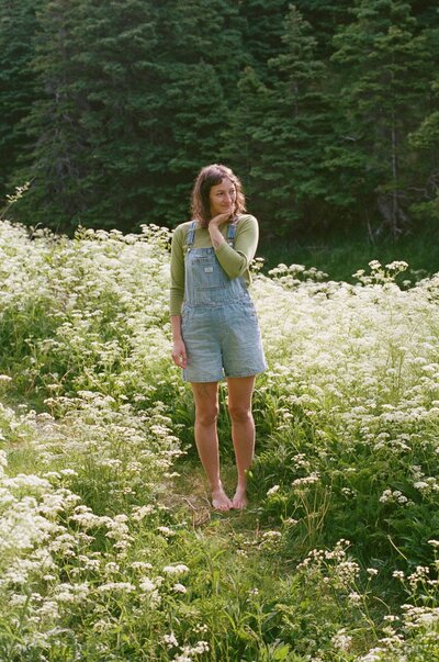 Heather giggles in a field of tall wildflowers. They wear overalls and a lime green shirt.