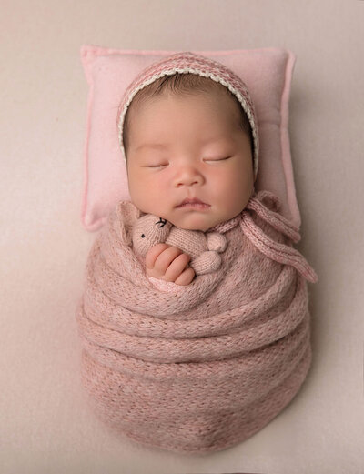 Newborn photography by Stamford CT photographer featuring baby girl in pink knit wrap with matching bonnet and tiny teddy bear prop