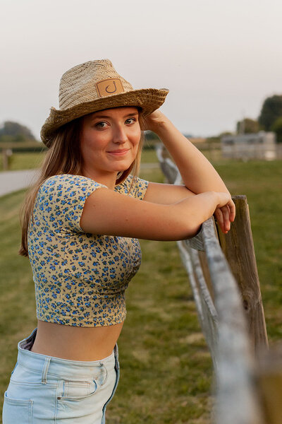 Image of senior girl facing a fence and leaning on it while holding onto the brim of her cowgirl hat and giving the camera a soft smile.