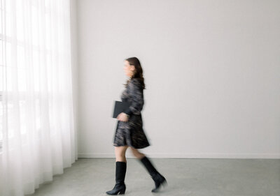 Out of focus business woman in dress and boots walking with laptop under her arms.
