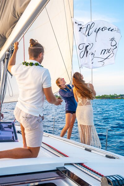 Couple raising a Marry Me flag on a sailboat in Hawaii as the man kneels to propose, with the ocean and coastline in the background.
