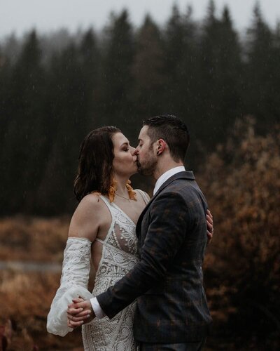 A boho wedding couple kissing in the rain with misty pine trees in the background.