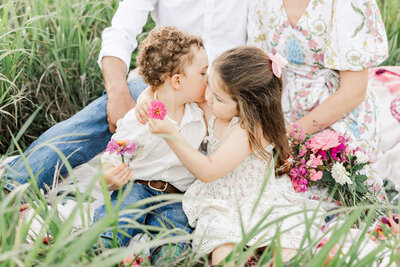 Little boy kissing his sister on the cheek, both of them holding a flower each while they sit in some long green grass.