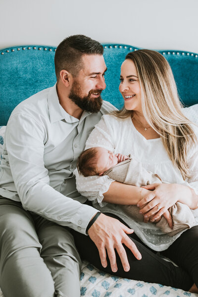 Newborn photo of mom and dad sitting next to each other on a bed holding newborn baby girl while wearing black and white in Davidsonville Maryland.