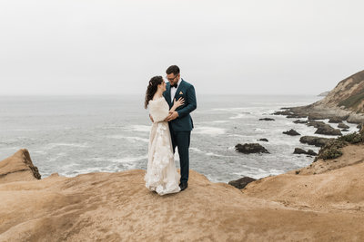 bride and groom standing on rock in front of ocean