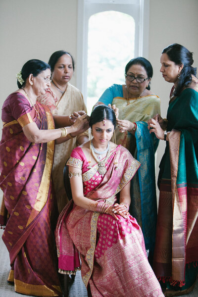 South Asian bride getting ready with women in traditional sarees inside a softly lit room, paired with a group portrait of a Western bride and bridesmaids in soft pink dresses and flower crowns, holding pastel bouquets. Wedding photography showcasing multicultural, emotional moments and luxury bridal styling.