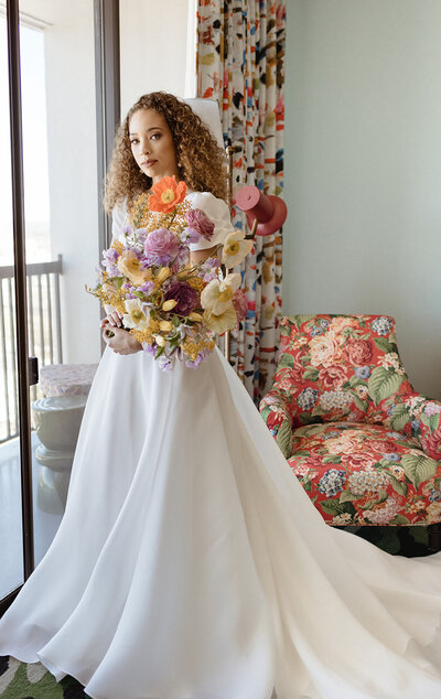 Modern bride in a flowing white gown holding a vibrant editorial-style bridal bouquet featuring orange poppies, yellow ranunculus, lavender lisianthus, buttercream tulips, and soft lilac blooms, photographed indoors against colorful patterned décor—luxury spring wedding floral design inspiration.