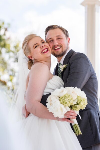 Bride and groom embrace hugging and laughing with their cheeks pressed together while bride holds white bouquet