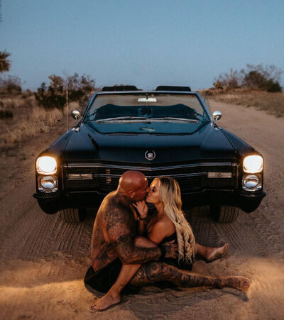 couple sitting in sand in front of car