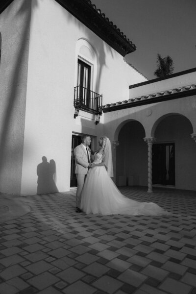 A bride and groom standing together in a tiled courtyard at night. The bride wears a ballgown with a long train, and they are framed by a white building with Spanish arches and a balcony.