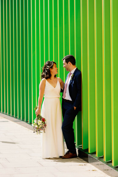 a newly married couple stood looking at each other in front of a colourful backdrop