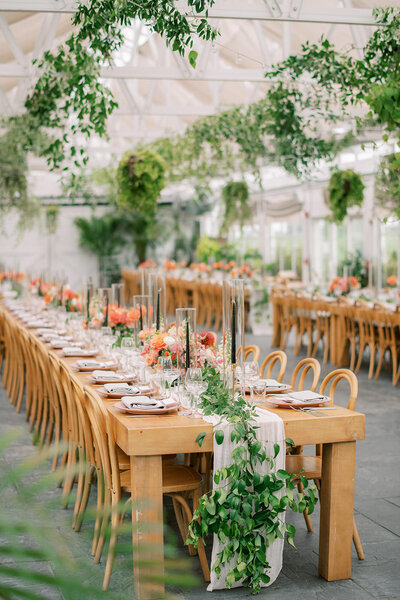 Wedding reception drenched in lush green botanicals. Bright white architectural details, long wooden farmhouse tables, black taper candles, and colorful florals feel like you're in a terrarium.