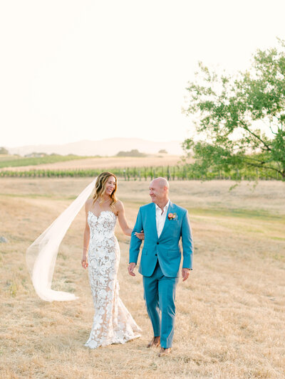 Bride and groom walk through field in Santa Ynez on their wedding day as veil blows in the breeze