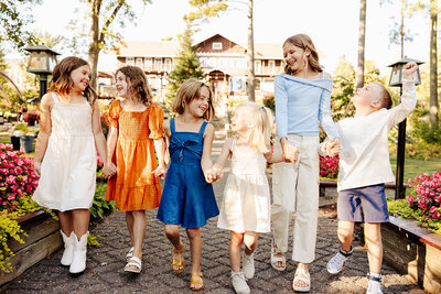 Six children happily walk on a path lined with flowers, their laughter echoing under the sunny sky. Dressed in colorful summer outfits and holding hands, they create an image of joy that Amber Bauer Photography beautifully captures as part of her Brainerd Lakes Area portfolio.
