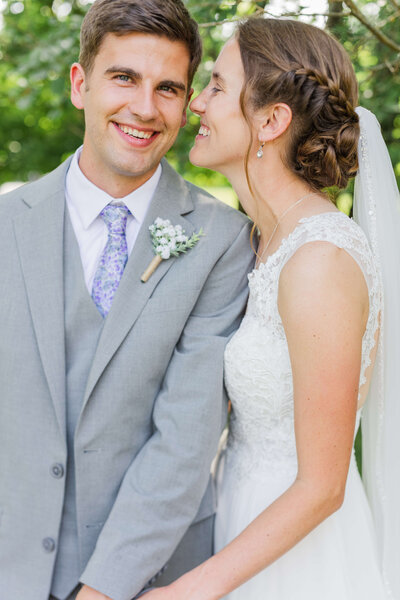 A groom in a grey suit smiles at the camera as his bride comes in close, smiling at him. She's looking at him as goes to put her hands on his hands.