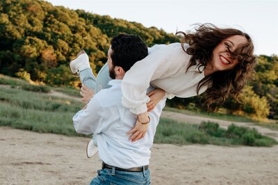 A playful and joyful engagement photo captured at Sunken Meadow Park in Long Island, NY. The groom-to-be, wearing a crisp white button-down shirt and jeans, lifts his fiancée over his shoulder as she laughs with her hair tousled in the breeze. She wears a flowing white blouse, light-wash ripped jeans, and white ankle boots, exuding a carefree and romantic energy. Her bright red lipstick and radiant smile contrast beautifully against the sunlit greenery in the background. The natural setting, with rolling hills and lush foliage, creates a dreamy, adventurous atmosphere, perfect for couples who embrace candid, fun-loving moments.
