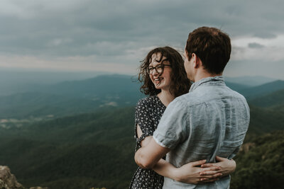 A woman with windswept hair hugs her husband on top of a mountain