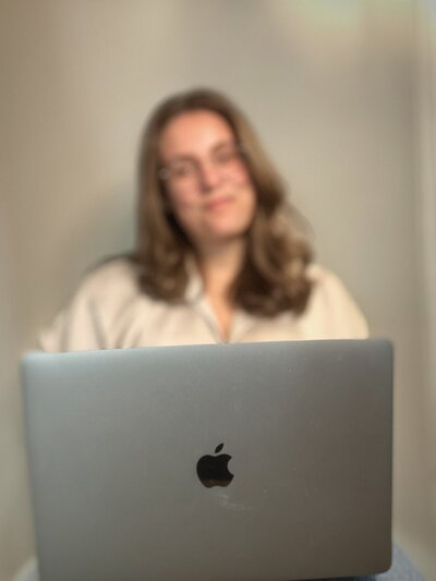 Woman with brown hair and glasses, smiling while sitting behind an open laptop, representing virtual assistant and web design services.