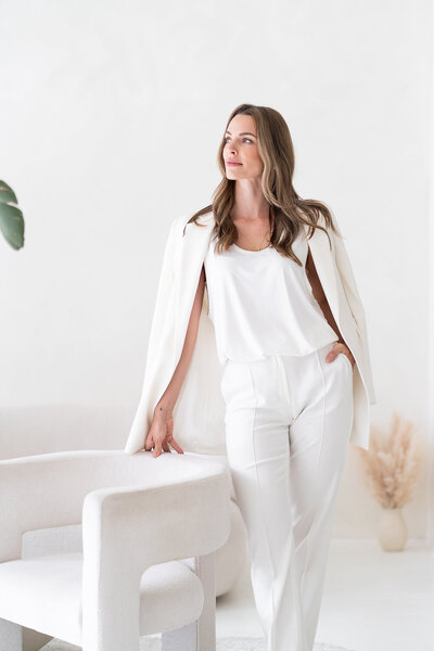 žena v bílém kostýmku stojí a rukou se opírá o bílé křeslo, dívá se doleva, za ní je bílá zeď, na zemi sušená tráva ve váze