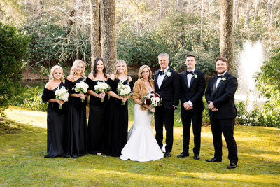 woman in white dress with fur coat holding flowers next to girls in black dresses and men in black tuxedos 