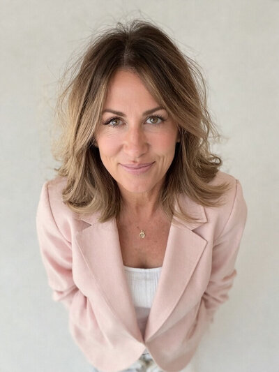 Woman with shoulder-length wavy hair, Cristina Horvath, CEO and brand strategist at Sweet Pea Ink Creative, wearing a light pink blazer and white top, stands and looks up at the camera against a neutral background.