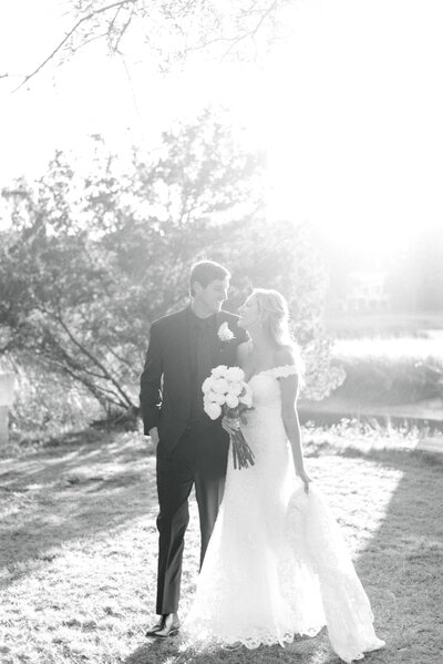 Bride and groom smiling at each other on black and white film bride with a white bouquet of long stem roses and groom in all black tux