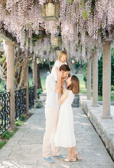 Young family with their child on dad’s shoulders sharing a sweet moment under blooming wisteria during a spring family photo session