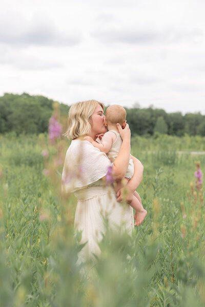 Mom holds her baby and kisses his cheek while both dressed in cream standing in a field with tall grass and flowers, Indianapolis Spring Mini Session