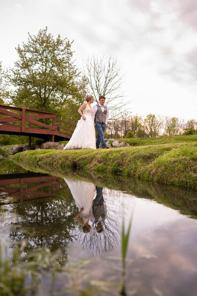 Bride and groom walk next to water with their reflection at sunset.
