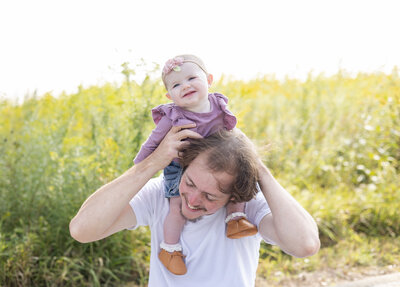 Father in a white t-shirt with his 6 mo old daughter wearing a purple shirt, blue jean shorts, brown shoes on his shoulders