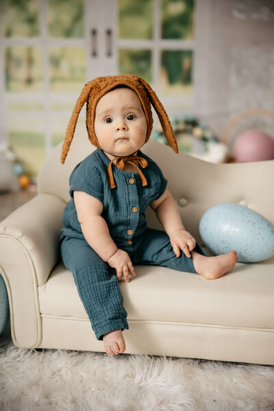 baby wearing a brown bunny bonnet, sitting on a small couch with blue and pink easter eggs. Studio photography