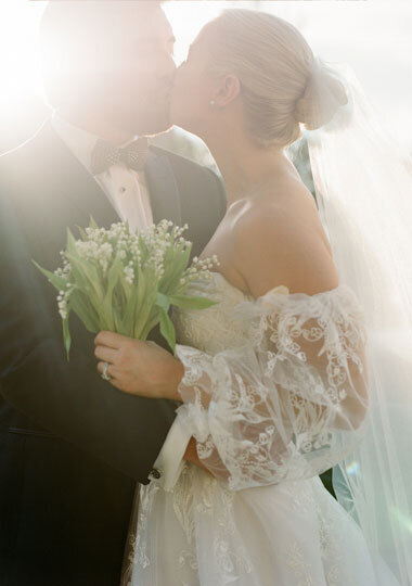 A bride and groom kiss in soft lighting. The bride wears an off-shoulder lace gown and holds a bouquet of lilies of the valley. The groom is in a black tuxedo.