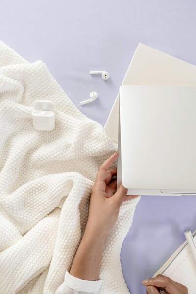 abriendo laptop, airpods con cobija blanca y fondo morado lilac