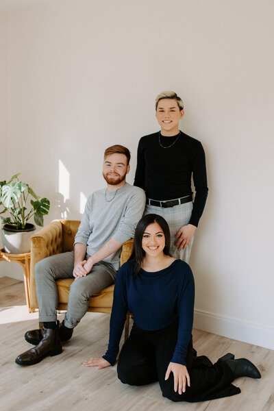 three people posing together. One is standing behind everyone, one is sitting in a chair, and one is sitting on the ground