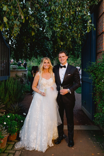 Bride and groom standing together in a romantic garden pathway, framed by lush greenery and elegant gate.