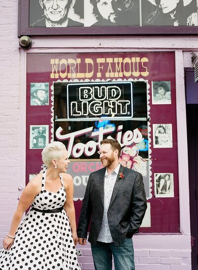 1950s pinup bride wearing a white dress with polk dots and a rockabilly groom wearing jeans a silk suit jacket and a red boutonniere smiling in front of Tootsies Honky Tonk on Broadway in Nashville